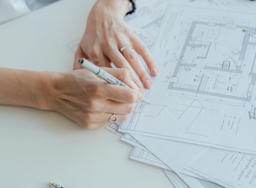 Person zeichnet auf Architektur- oder Baupl&auml;nen, Skizzenblock und Stifte auf wei&szlig;em Tisch, Planungsarbeit im B&uuml;ro.