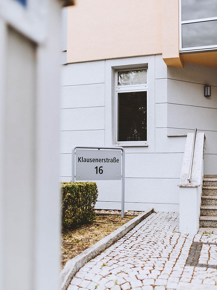Eingangsbereich eines Wohnhauses in der Klausenerstraße 16 mit Hausnummernschild, gepflastertem Weg und Treppe.