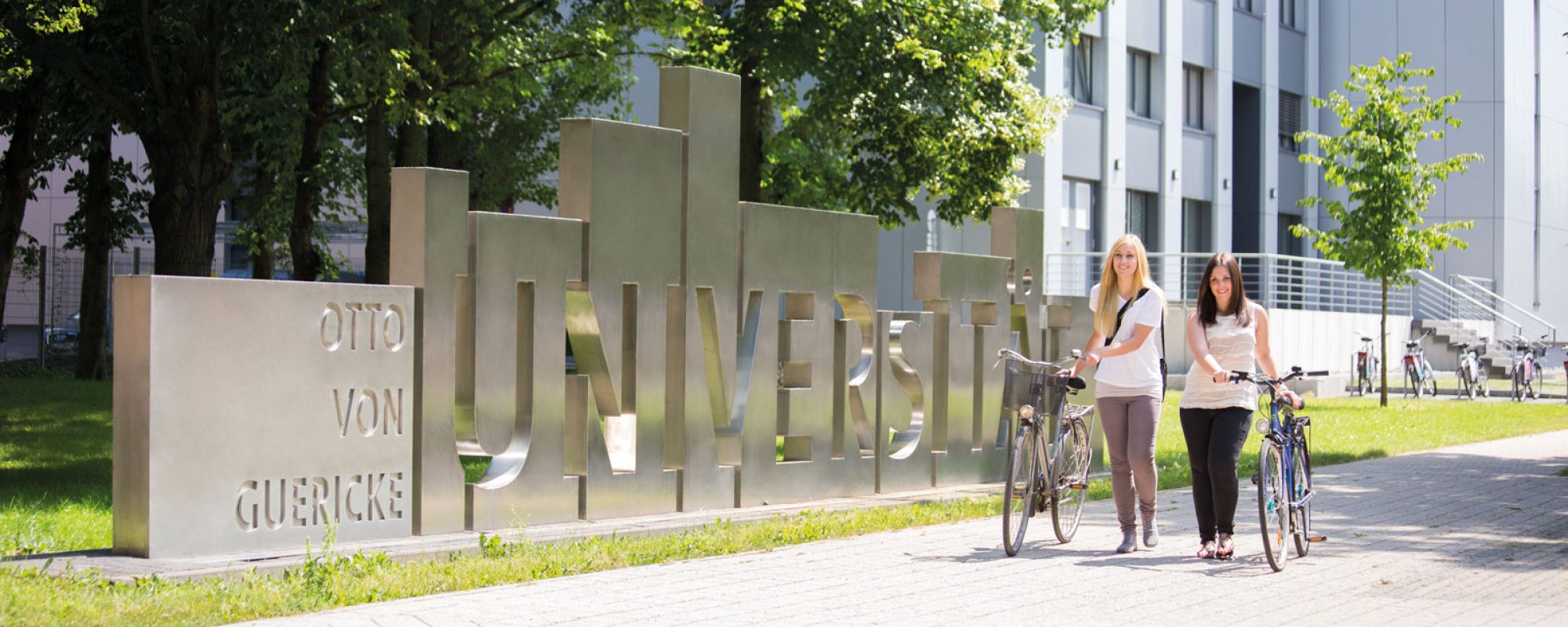 Otto-von-Guericke-Universität Magdeburg, Studierende mit Fahrrädern vor Uniskulptur auf grünem Campus.